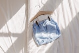 A pair of light blue shorts with a subtle dot pattern is clipped to a wooden hanger. The shorts are placed on a soft, sunlit bedspread, creating a calm and serene atmosphere.