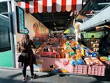 A woman stands in front of a vibrant candy store. The store displays a wide variety of sweets, including colorful lollipops, candy canes, and an assortment of chips in brightly colored bags. The shop has a playful, inviting atmosphere with its red-and-white striped awning and decorative signage.