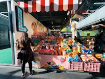 A woman stands in front of a vibrant candy store. The store displays a wide variety of sweets, including colorful lollipops, candy canes, and an assortment of chips in brightly colored bags. The shop has a playful, inviting atmosphere with its red-and-white striped awning and decorative signage.