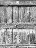 Weathered wooden planks are aligned vertically, fastened with square metal bolts. The texture of the wood shows visible grain patterns and signs of aging, such as discoloration and cracks.