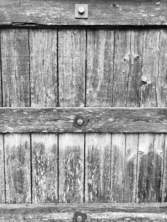 Weathered wooden planks are aligned vertically, fastened with square metal bolts. The texture of the wood shows visible grain patterns and signs of aging, such as discoloration and cracks.