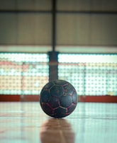 A black soccer ball with red seams sits on a wooden gymnasium floor. The background features large, blurred windows with a grid-like pattern, suggesting an indoor sports venue.