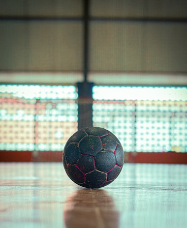 A black soccer ball with red seams sits on a wooden gymnasium floor. The background features large, blurred windows with a grid-like pattern, suggesting an indoor sports venue.