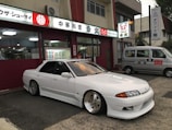 A sleek white sports car with custom modifications is parked in front of a traditional Japanese restaurant with red and white signage. The restaurant's signage contains Japanese characters and symbols, giving a cultural context. Next to the car, a silver van is parked, displaying similar symbols and writing.