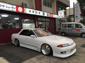 A sleek white sports car with custom modifications is parked in front of a traditional Japanese restaurant with red and white signage. The restaurant's signage contains Japanese characters and symbols, giving a cultural context. Next to the car, a silver van is parked, displaying similar symbols and writing.
