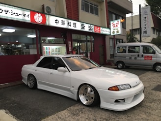 A sleek white sports car with custom modifications is parked in front of a traditional Japanese restaurant with red and white signage. The restaurant's signage contains Japanese characters and symbols, giving a cultural context. Next to the car, a silver van is parked, displaying similar symbols and writing.