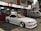 A sleek white sports car with custom modifications is parked in front of a traditional Japanese restaurant with red and white signage. The restaurant's signage contains Japanese characters and symbols, giving a cultural context. Next to the car, a silver van is parked, displaying similar symbols and writing.