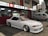 A sleek white sports car with custom modifications is parked in front of a traditional Japanese restaurant with red and white signage. The restaurant's signage contains Japanese characters and symbols, giving a cultural context. Next to the car, a silver van is parked, displaying similar symbols and writing.