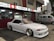 A sleek white sports car with custom modifications is parked in front of a traditional Japanese restaurant with red and white signage. The restaurant's signage contains Japanese characters and symbols, giving a cultural context. Next to the car, a silver van is parked, displaying similar symbols and writing.