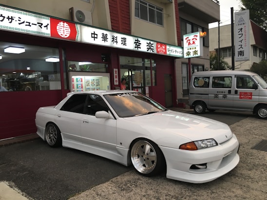 A sleek white sports car with custom modifications is parked in front of a traditional Japanese restaurant with red and white signage. The restaurant's signage contains Japanese characters and symbols, giving a cultural context. Next to the car, a silver van is parked, displaying similar symbols and writing.