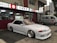 A sleek white sports car with custom modifications is parked in front of a traditional Japanese restaurant with red and white signage. The restaurant's signage contains Japanese characters and symbols, giving a cultural context. Next to the car, a silver van is parked, displaying similar symbols and writing.