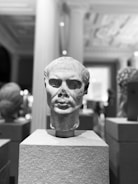 A monochrome photograph showcases an ancient stone bust displayed on a pedestal in a museum setting. The background reveals blurred outlines of other sculptures and elements of classical architecture like columns.