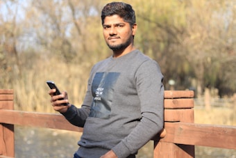 A person stands leaning on a wooden fence holding a smartphone in one hand, outdoors in a natural setting with trees and a blurred background. They are wearing a gray long-sleeved shirt with a design on the front. The expression appears calm and relaxed.
