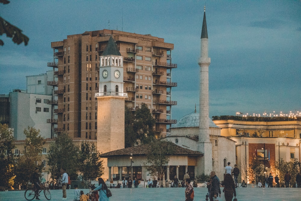 a group of people walking around a plaza with a clock tower in the background