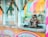 A fluffy, long-haired cat sits on the counter of a colorful food stand. The stand features bright signage with playful, pastel colors. The cat seems to be gazing out, appearing calm and composed amidst the vibrant setting.