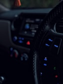 A close-up view of a car's interior, focusing on the steering wheel with visible control buttons such as volume and mute. The dashboard is dimly lit, showing blurred controls and displays with prominent use of blue and red lights.