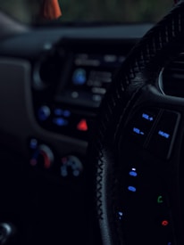 A close-up view of a car's interior, focusing on the steering wheel with visible control buttons such as volume and mute. The dashboard is dimly lit, showing blurred controls and displays with prominent use of blue and red lights.