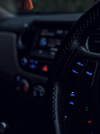 A close-up view of a car's interior, focusing on the steering wheel with visible control buttons such as volume and mute. The dashboard is dimly lit, showing blurred controls and displays with prominent use of blue and red lights.