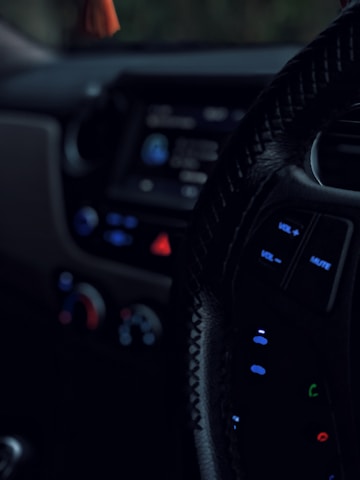 A close-up view of a car's interior, focusing on the steering wheel with visible control buttons such as volume and mute. The dashboard is dimly lit, showing blurred controls and displays with prominent use of blue and red lights.