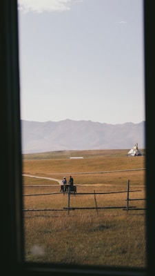 A serene landscape view with two people riding horses on a path in an open grassy field. A wooden fence runs across the foreground, and in the background, a mountainous range is visible under a clear sky. A solitary tent or tipi can be seen in the distance.