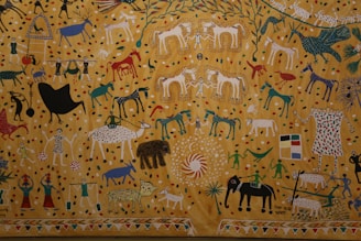 A vibrant and detailed painting featuring various animals, people, and abstract shapes. The background consists of a golden yellow with colorful dots scattered throughout. The animals depicted include horses, elephants, tigers, and birds, all rendered in a stylized and colorful manner. There are also human figures dressed in traditional attire participating in activities like celebration and carrying objects. The scene is lively and full of movement, with intricate patterns and symbols decorating the elements.