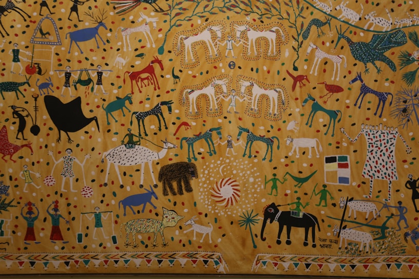 A vibrant and detailed painting featuring various animals, people, and abstract shapes. The background consists of a golden yellow with colorful dots scattered throughout. The animals depicted include horses, elephants, tigers, and birds, all rendered in a stylized and colorful manner. There are also human figures dressed in traditional attire participating in activities like celebration and carrying objects. The scene is lively and full of movement, with intricate patterns and symbols decorating the elements.