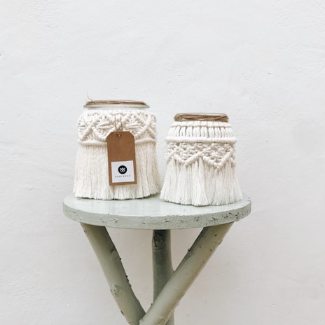 Two intricately designed macrame jars sit on a small, rustic wooden table. The jars are adorned with cream-colored woven patterns and fringes, with one having a brown tag attached. The background is a plain, light-colored wall, adding to the minimalist aesthetic.