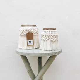 Two intricately designed macrame jars sit on a small, rustic wooden table. The jars are adorned with cream-colored woven patterns and fringes, with one having a brown tag attached. The background is a plain, light-colored wall, adding to the minimalist aesthetic.