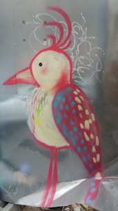 A colorful, stylized bird painted on a metallic surface with abstract graffiti patterns in the background. The bird features a bright red outline with a yellow and white body, blue and red spotted wings, and three feathery tufts on its head.