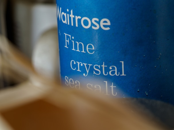 Close-up of a blue container labeled 'Waitrose Fine crystal sea salt,' with slightly blurred elements in the foreground creating a sense of depth.