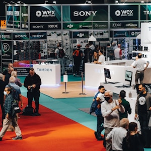 A busy exhibition booth featuring brands like Sony, Tamron, and DJI, where several people are interacting with product displays and browsing. The booth is well-lit with overhead lighting, and signs promote camera equipment and technology.