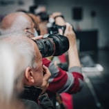 A group of photographers holding cameras with large lenses, aiming in the same direction. The focus is on one photographer in the foreground, while others appear blurred in the background.