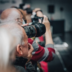 A group of photographers holding cameras with large lenses, aiming in the same direction. The focus is on one photographer in the foreground, while others appear blurred in the background.