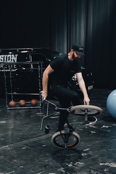 A man performs a bicycle trick indoors on a dark stage. He is balancing on the front wheel of a BMX bike, wearing black clothing and a cap. In the background, there are three basketballs on a rack, a blue exercise ball, and a black van with branding on the side.