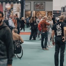 People are gathered in a busy exhibition hall or convention center, some appear to be in conversation while others are browsing displays or products. The atmosphere is lively, with a variety of individuals, some carrying backpacks and others in casual attire.