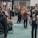 People are gathered in a busy exhibition hall or convention center, some appear to be in conversation while others are browsing displays or products. The atmosphere is lively, with a variety of individuals, some carrying backpacks and others in casual attire.