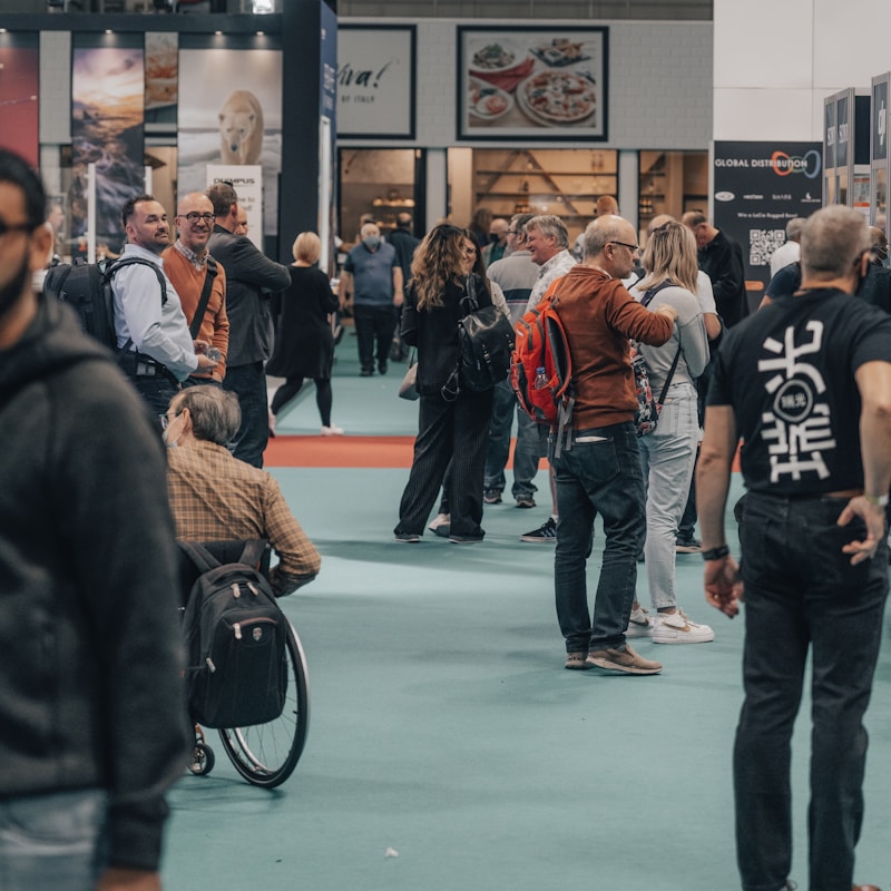 People are gathered in a busy exhibition hall or convention center, some appear to be in conversation while others are browsing displays or products. The atmosphere is lively, with a variety of individuals, some carrying backpacks and others in casual attire.