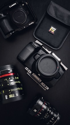 A variety of professional camera equipment includes a Lumix camera with protective casing, a DSLR camera body, and several camera lenses. A padded case with an FX logo is also visible. The equipment is arranged on a dark surface, showcasing the gear's sleek and robust design.