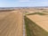 An aerial view of vast agricultural fields stretching to the horizon. The landscape is divided into several sections with different types of crops, flanked by a long, straight road lined with trees. In the distance, industrial buildings and mountains can be seen under a clear blue sky.