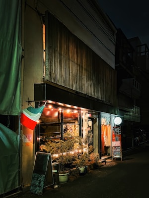 A cozy, dimly lit storefront with a variety of potted plants outside. The building has a small awning, and strings of warm lights illuminate the entrance. An Italian flag hangs beside the door. Menus or signs are displayed on a chalkboard and stand, and vibrant orange curtains can be seen inside.