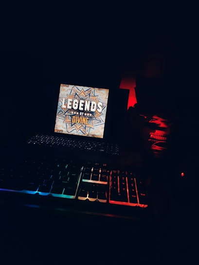 A dark environment featuring a brightly lit screen with a decorative design and the words 'LEGENDS BY DIVINE.' Below the screen, an illuminated mechanical keyboard displays rainbow lighting. The scene is dimly lit with some red ambient light in the background.