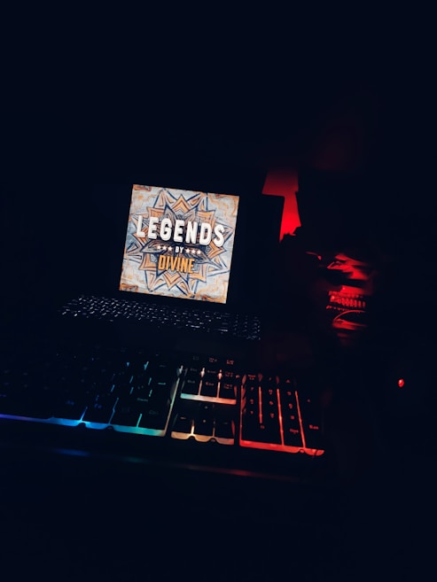 A dark environment featuring a brightly lit screen with a decorative design and the words 'LEGENDS BY DIVINE.' Below the screen, an illuminated mechanical keyboard displays rainbow lighting. The scene is dimly lit with some red ambient light in the background.