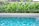 A clear swimming pool with vibrant blue tiles is set in the foreground, surrounded by lush green tropical plants. The pool water reflects the intricate patterns of the tiles, creating a serene and inviting atmosphere.