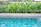 A clear swimming pool with vibrant blue tiles is set in the foreground, surrounded by lush green tropical plants. The pool water reflects the intricate patterns of the tiles, creating a serene and inviting atmosphere.