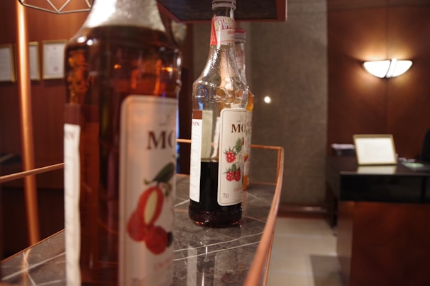 A close-up of liquor bottles with one prominently labeled bottle featuring berry illustrations. The bottles are displayed on a sleek, dark marble surface. The background includes a warmly lit interior with wood paneling and a modern lamp, along with some framed documents on the wall.