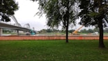 A construction site near a bridge with visible machinery including an excavator and a crane. Trees frame the image, and an orange safety fence separates the green grass area from the construction zone. The sky is overcast.