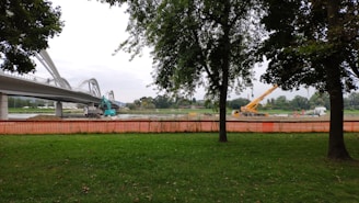 A construction site near a bridge with visible machinery including an excavator and a crane. Trees frame the image, and an orange safety fence separates the green grass area from the construction zone. The sky is overcast.