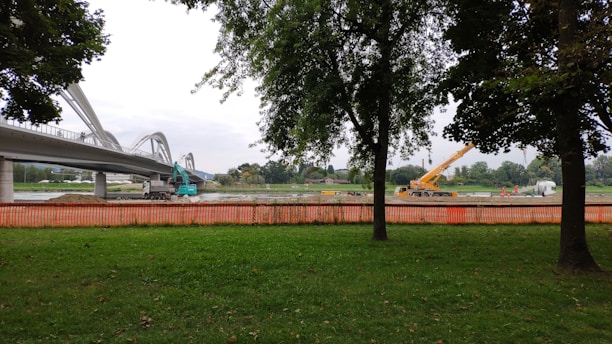 A construction site near a bridge with visible machinery including an excavator and a crane. Trees frame the image, and an orange safety fence separates the green grass area from the construction zone. The sky is overcast.