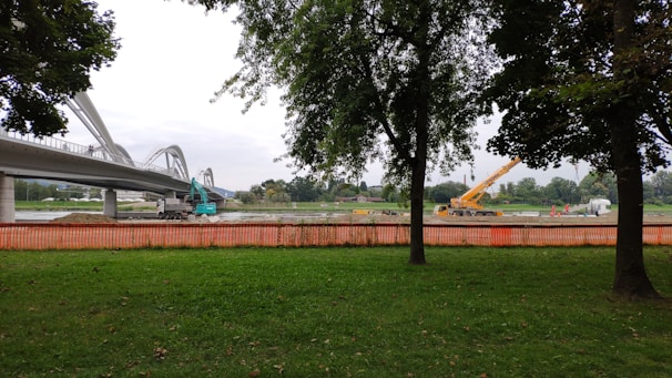 A construction site near a bridge with visible machinery including an excavator and a crane. Trees frame the image, and an orange safety fence separates the green grass area from the construction zone. The sky is overcast.