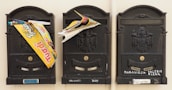Three black mailboxes are mounted on a wall. Each mailbox displays a decorative emblem. Printed materials and advertisements are stuffed in the gaps of two mailboxes. Names are written on the bottom of each mailbox.